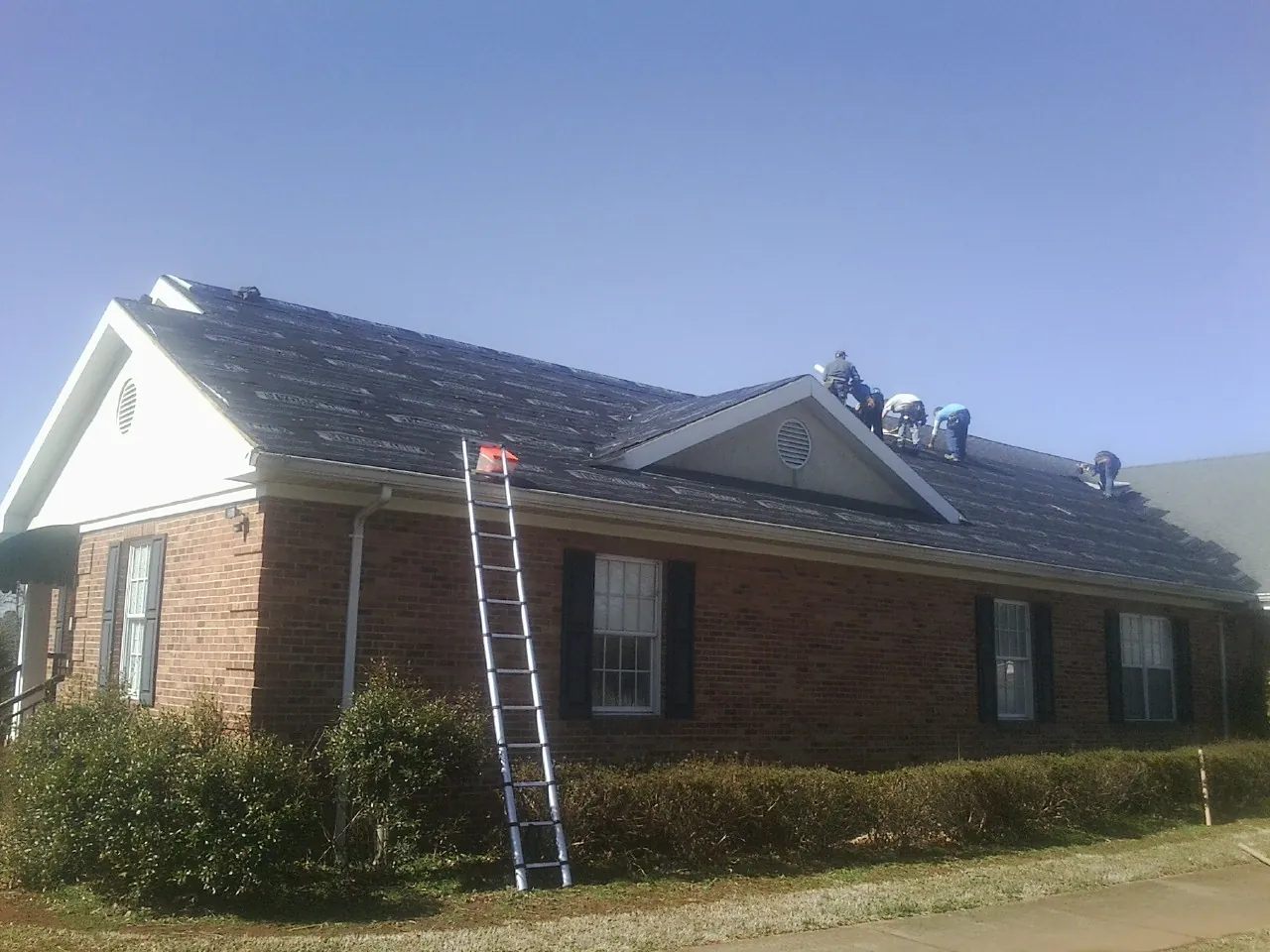 Crew tearing shingles off a brick ranch roof with a ladder set — Emergency Roof Repair work by our crew in Beach Lake