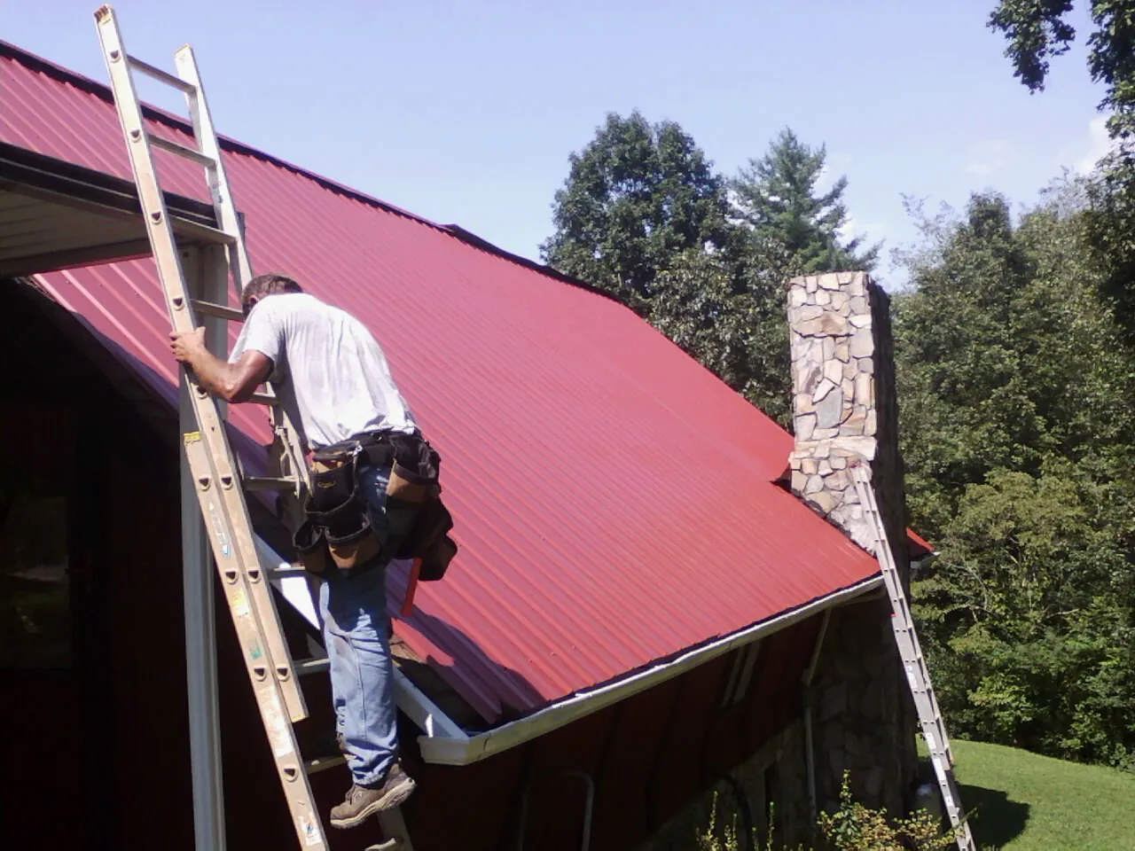 Roofer on a ladder beside a red metal roof and stone chimney — Soffit & Fascia Repair by Roofing Brothers in Beach Lake